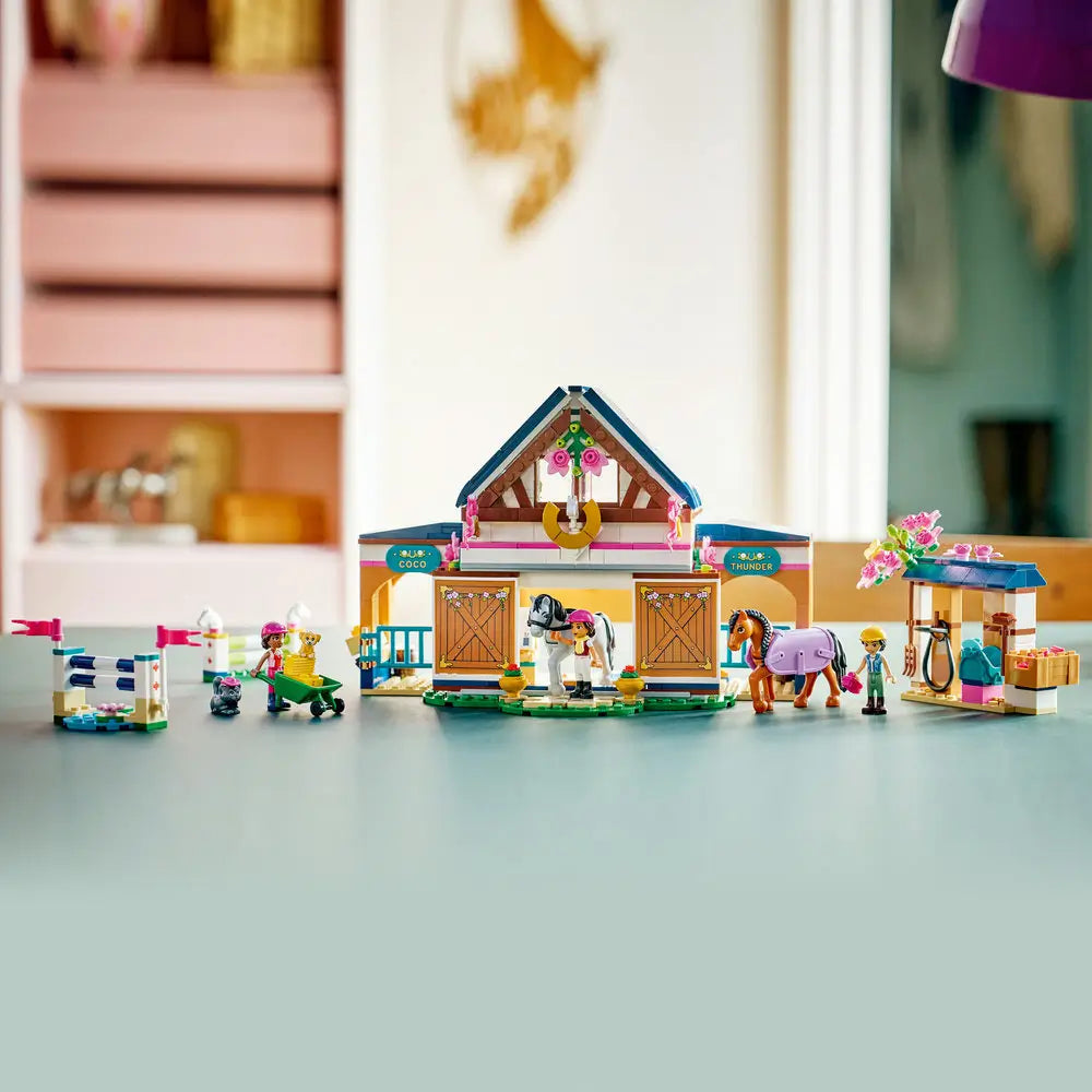 LEGO Friends Horse Stable and Riding Academy displayed model shows complete tan, white, and blue plastic barn with arches, stalls, flower accents, horses, jumps, mini dolls, and stable accessories indoors.