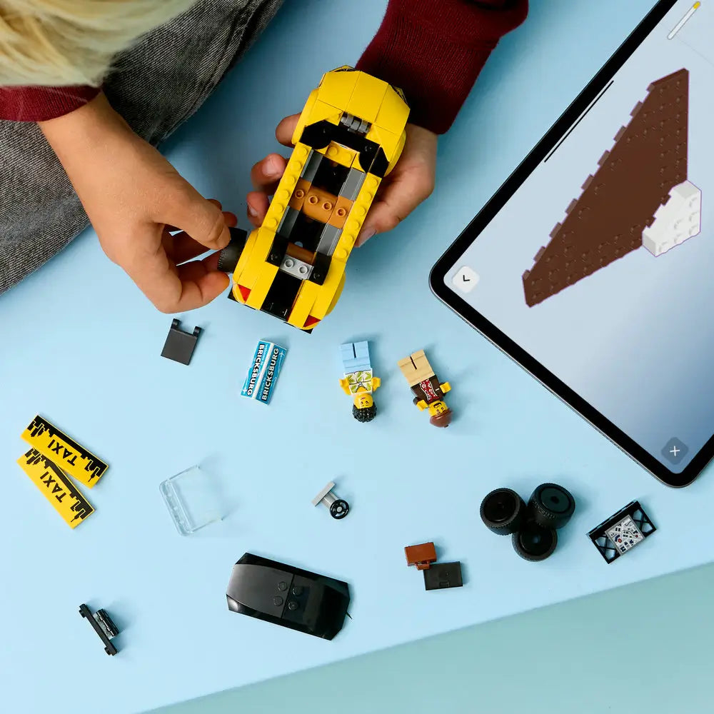 LEGO City taxi building process showing hands assembling a yellow brick car with loose pieces, minifigures, wheels, and instructions nearby, illustrating creative construction play on a light blue surface.