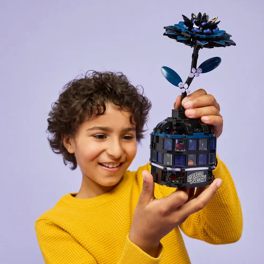 Smiling child holds up the completed LEGO Black Dahlia Flower set from Wednesday, featuring a tall dark flower emerging from a black glass dome structure with visible interior details and a Nevermore Academy label.