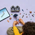 Overhead view of child assembling the LEGO Black Dahlia Flower set from Wednesday, surrounded by colorful bricks, mini-dolls, accessories, and a tablet displaying digital building instructions.
