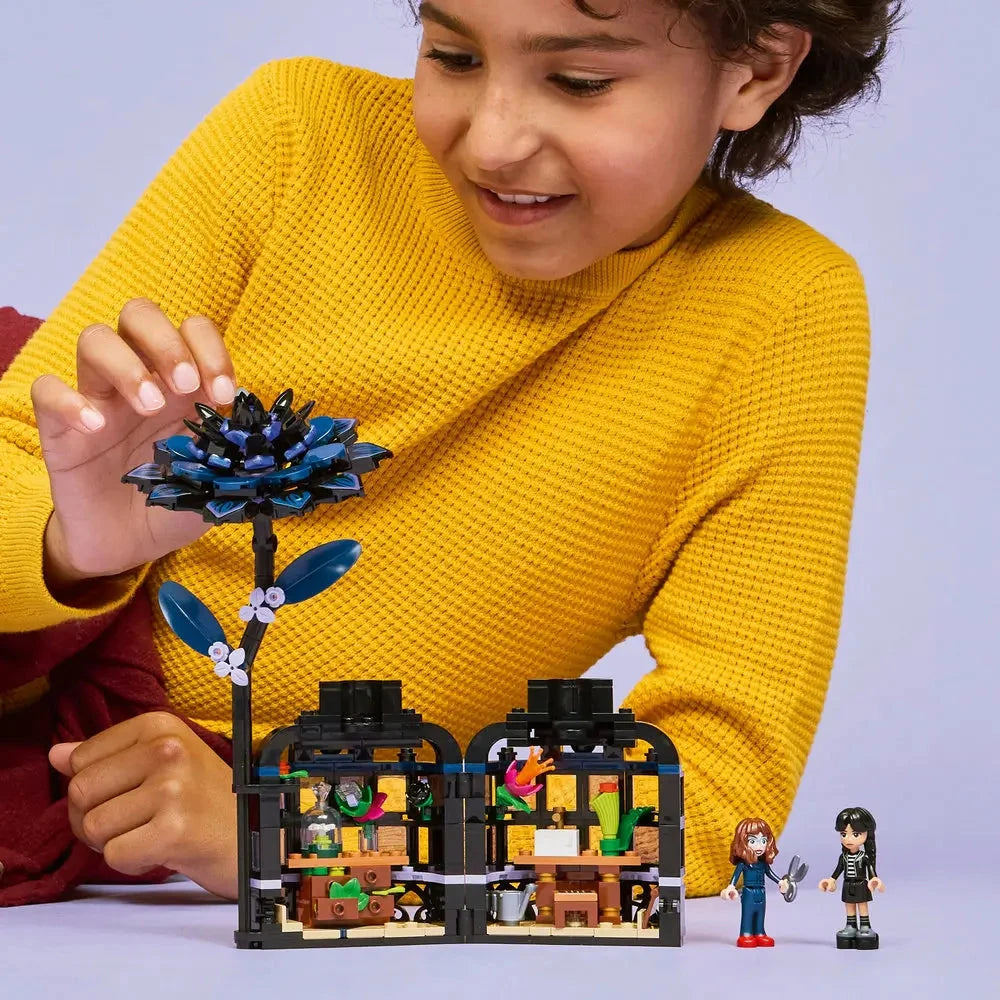 Smiling child in yellow sweater plays with the LEGO Black Dahlia Flower set from Wednesday, showing the tall dark flower connected to an open buildable greenhouse with vibrant plant details and mini-dolls.