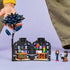A child’s hand interacts with a LEGO Wednesday Addams Black Dahlia Flower set, showing a dark blue and black buildable flower atop a detailed room scene with two mini-dolls and colorful botanical accessories.
