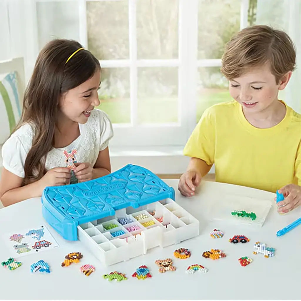 Aquabeads Beginners Craft Studio craft activity with two children using blue bead case and peg board to create colorful animal and vehicle bead designs at a white table indoors.