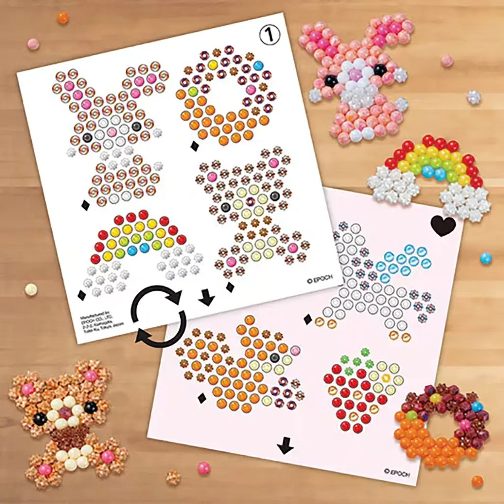 Aquabeads Beginners Craft Studio pattern sheets displaying printed bead placement guides for bunny, donut, rainbow, fox, and strawberry designs on a wooden tabletop.