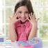 Smiling child wearing a pink outfit showing off her decorated nails after using the Aquabeads Nail Studio, with crafting trays and colorful nail accessories visible on the table.