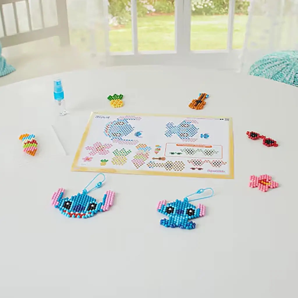 Tabletop view of Disney Aquabeads Stitch craft setup showing a bead design template, spray bottle, and completed bead figures including Stitch, sunglasses, flower, and ice cream cone.