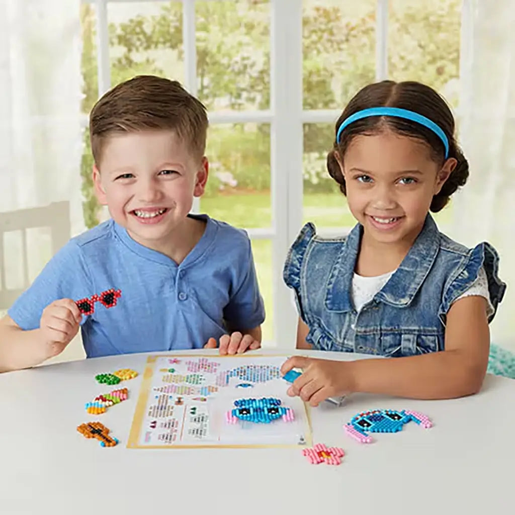 Two children at a white table smiling as they create Disney Aquabeads Stitch designs, surrounded by colorful bead pieces and a crafting guide in a bright indoor playroom.