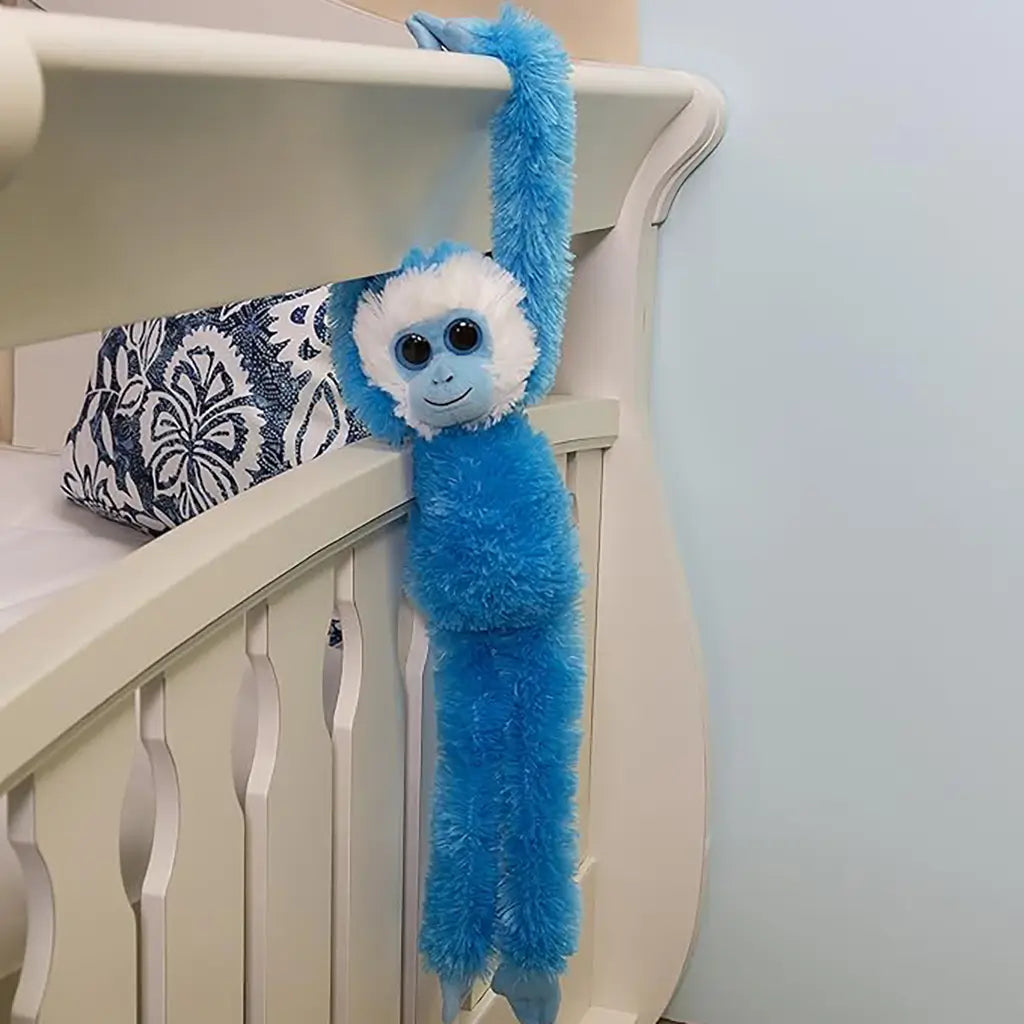 Aurora hanging monkey plush toy with vivid blue soft polyester fur, long flexible arms gripping furniture, slim dangling body, and cheerful face, styled as playful room decor in a nursery environment.