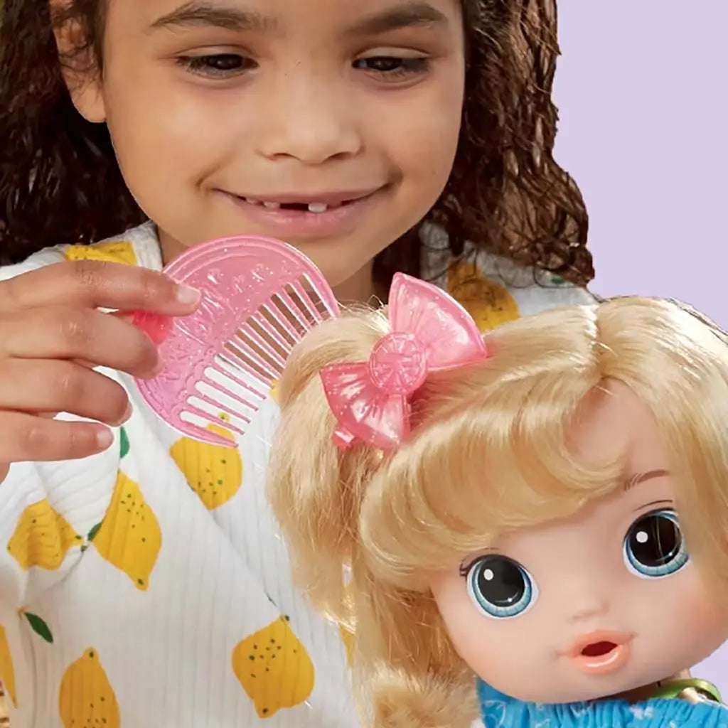 Child brushing Baby Alive doll’s blonde hair using a pink comb, highlighting the doll’s bright eyes, bow accessory, and colorful dress in a playful scene.