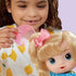 Child brushing Baby Alive doll’s blonde hair using a pink comb, highlighting the doll’s bright eyes, bow accessory, and colorful dress in a playful scene.