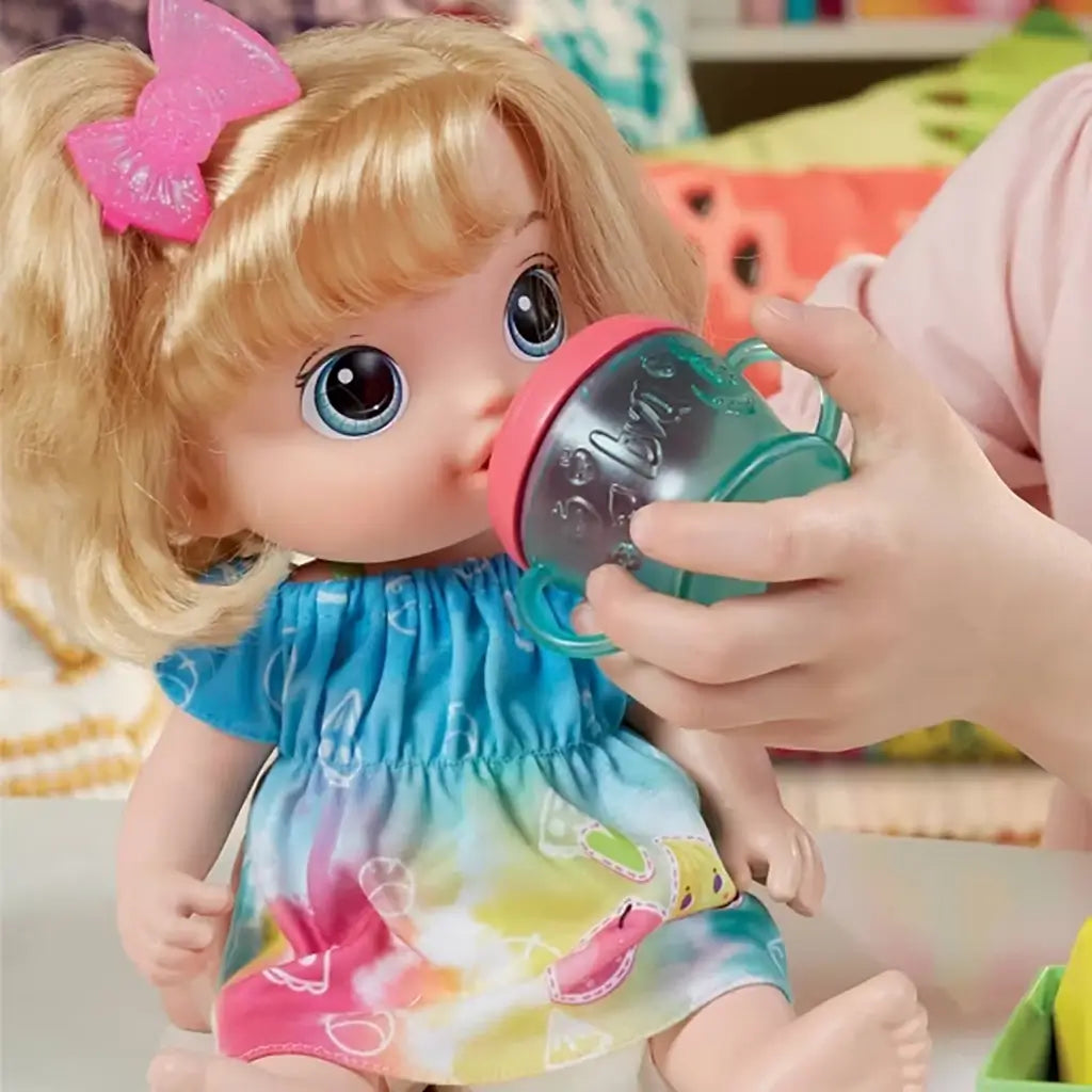 Close-up of Baby Alive doll being fed with teal sippy cup, showing its wide blue eyes, blonde bow, and colorful dress during imaginative feeding play.