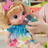 Close-up of Baby Alive doll being fed with teal sippy cup, showing its wide blue eyes, blonde bow, and colorful dress during imaginative feeding play.