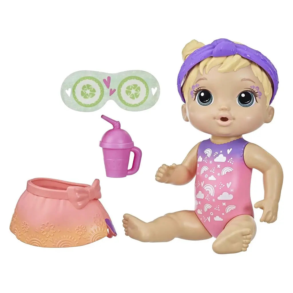 Baby Alive spa doll shows peach-tone plastic figure in a pink-to-purple rainbow swimsuit with a purple headband, seated beside a peach skirt, pink sippy cup, and cucumber-print eye mask arranged neatly on a white backdrop.