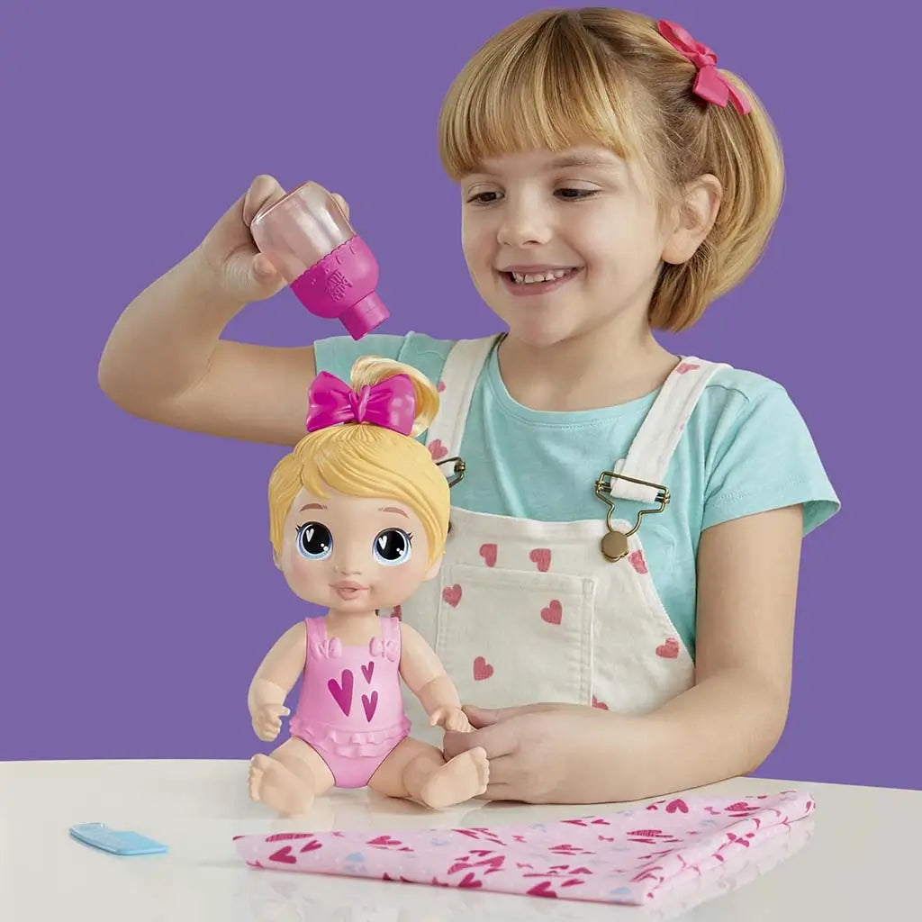 Child happily feeding Baby Alive Harper Hugs doll using pink bubble bottle, with blond doll in pink heart outfit seated on table beside blanket for nurturing pretend play.