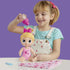 Child happily feeding Baby Alive Harper Hugs doll using pink bubble bottle, with blond doll in pink heart outfit seated on table beside blanket for nurturing pretend play.