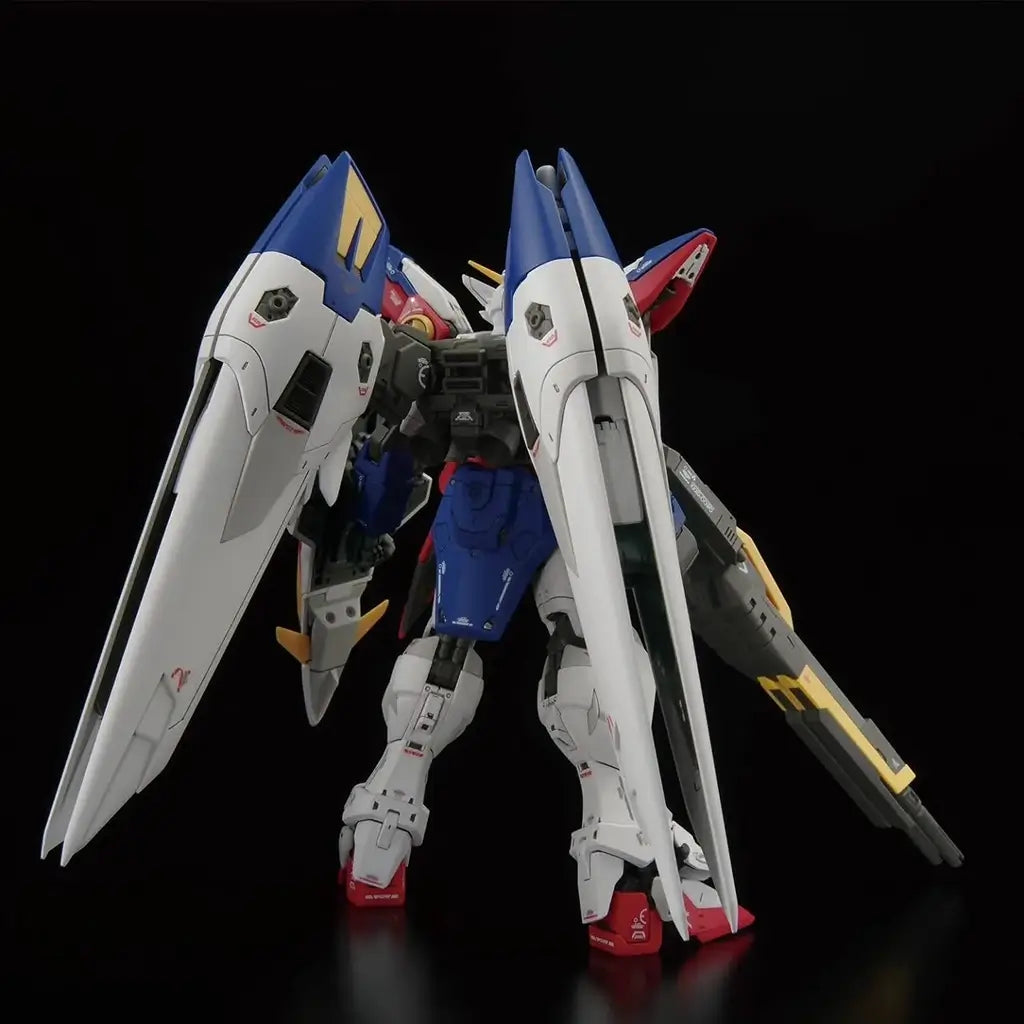 Bandai RG Wing Gundam Zero model kit rear view reveals the mechanical detailing inside the large white and blue wing units, backpack frame and leg armor, with the long buster rifle resting at the mobile suit’s side.