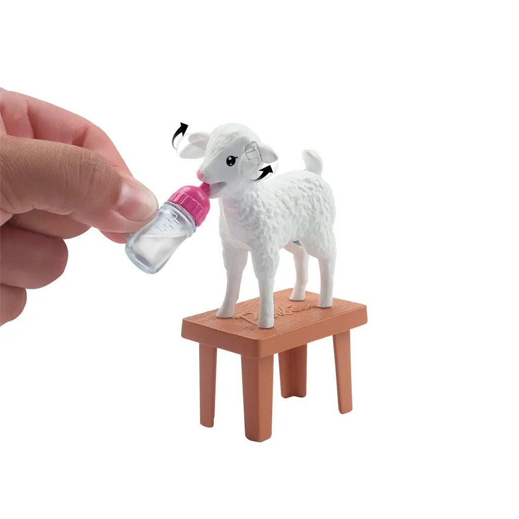 Close-up of white lamb toy from Barbie farm vet set standing on brown exam table, being bottle-fed with pink baby bottle accessory for pretend veterinary care.