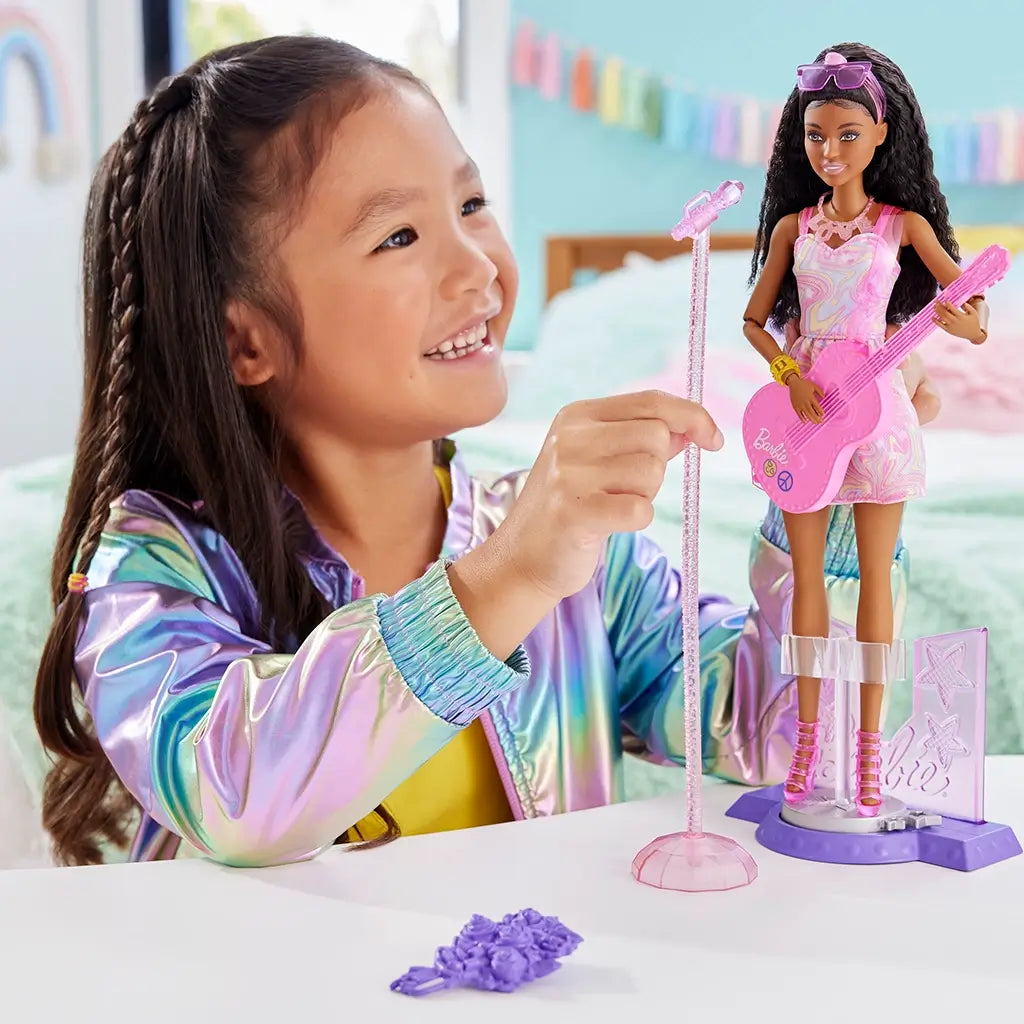 Barbie 65th Anniversary Pop Star Doll set shows a smiling child in a rainbow jacket playing with the doll dressed in a pastel swirl dress, pink guitar, and stage microphone.