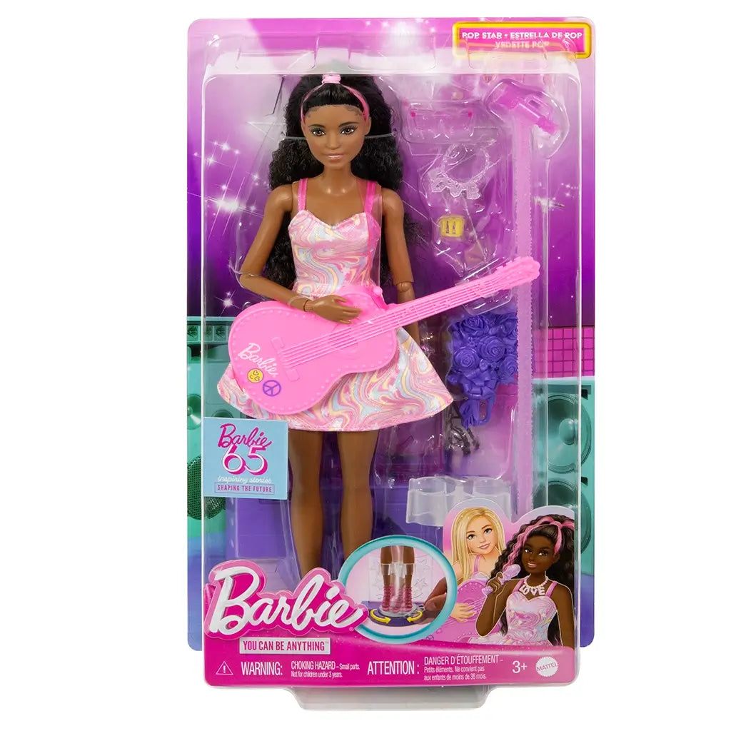 Barbie 65th Anniversary Pop Star Doll in retail packaging, featuring the doll with pink guitar, pastel swirl dress, and included stage accessories displayed against a purple backdrop.