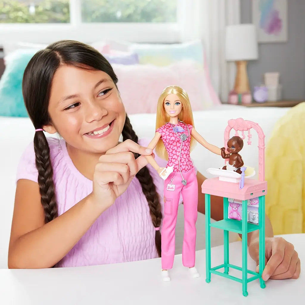 Smiling young girl plays with Barbie baby doctor doll dressed in pink scrubs and nursery station, engaging with infant doll in diaper for realistic medical role-play fun.