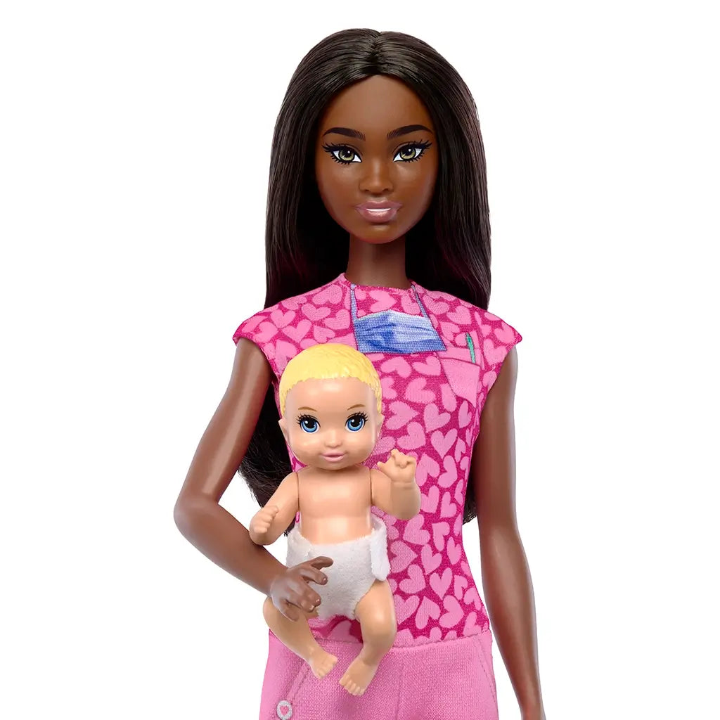 Barbie Baby Doctor doll with long dark hair wears pink scrubs patterned with hearts while holding a blonde baby doll in a diaper, highlighting her nurturing pediatric role.