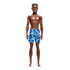 Front view of Barbie Beach Ken doll standing barefoot, wearing blue camouflage swim shorts, with short brown textured hair and a smiling facial expression.