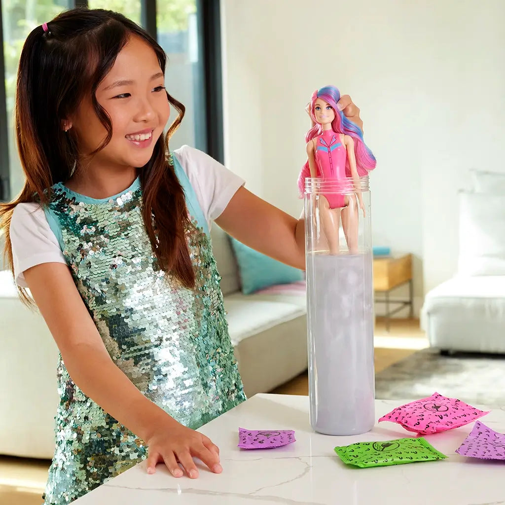 Young girl in a shiny teal sequin dress smiles while lifting a Barbie Color Reveal doll with pink and blue hair from a tall water-filled tube on a white kitchen counter.