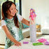 Young girl in a shiny teal sequin dress smiles while lifting a Barbie Color Reveal doll with pink and blue hair from a tall water-filled tube on a white kitchen counter.