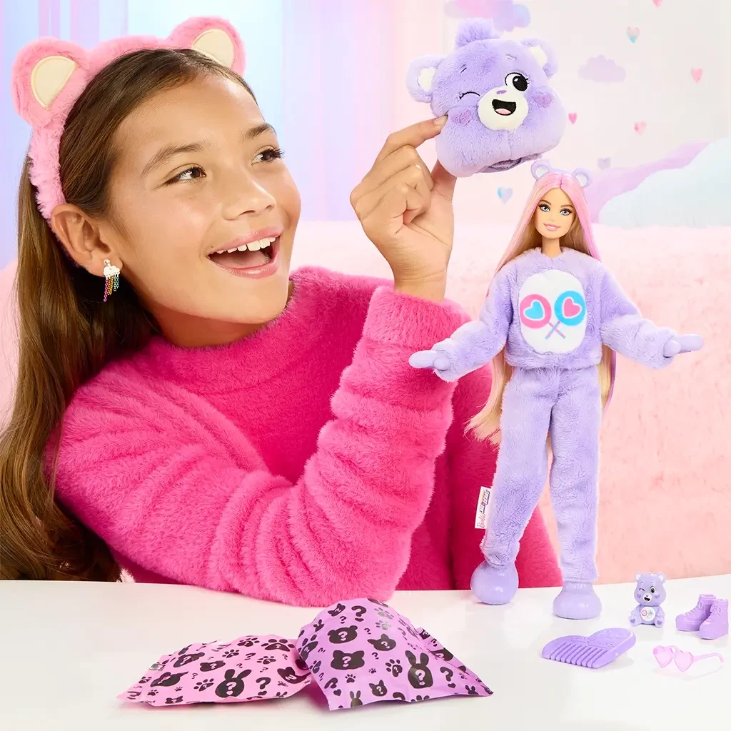 Smiling girl holding a plush purple bear head while playing with Barbie dressed in matching costume, with accessories and surprise bags on the table.