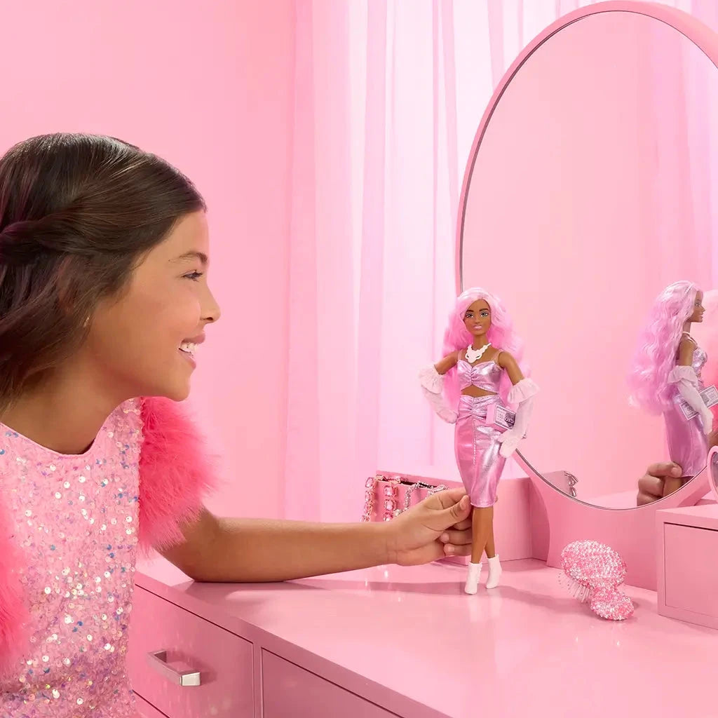 Smiling child plays with Barbie Deluxe doll dressed in a shiny pink ruched dress, white boots, pink hair, and matching gloves, standing in front of a vanity.