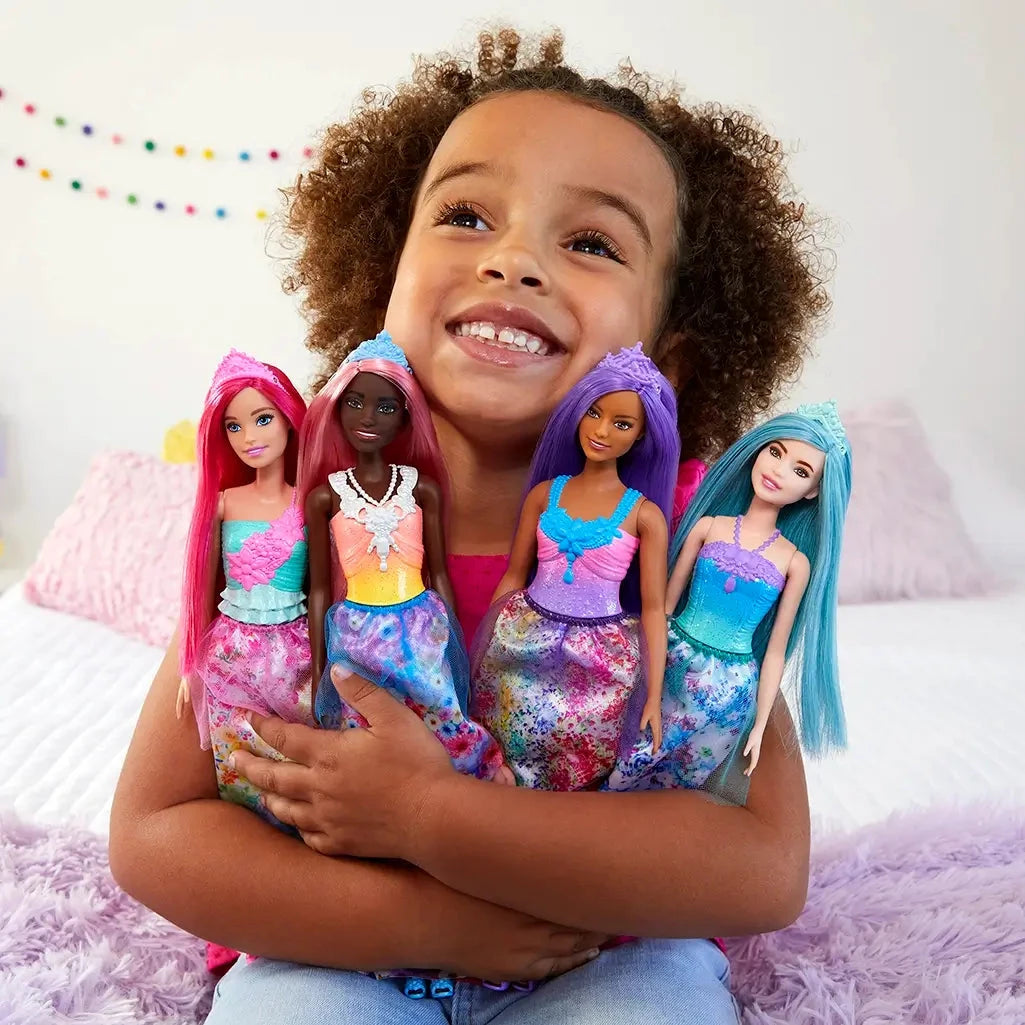 Smiling child hugging four Barbie Dreamtopia princess dolls, each with colorful hair, tiaras, and fantasy-themed gowns, on a cozy bed with rainbow decor.
