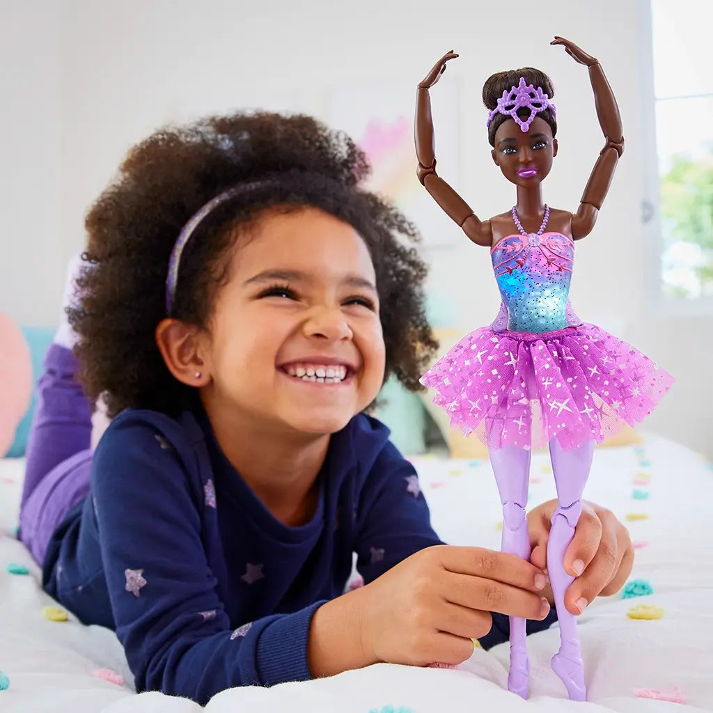 Smiling young girl plays with Barbie Dreamtopia ballerina doll, dressed in a glittery pink tutu and lavender tights, with light-up bodice and tiara.