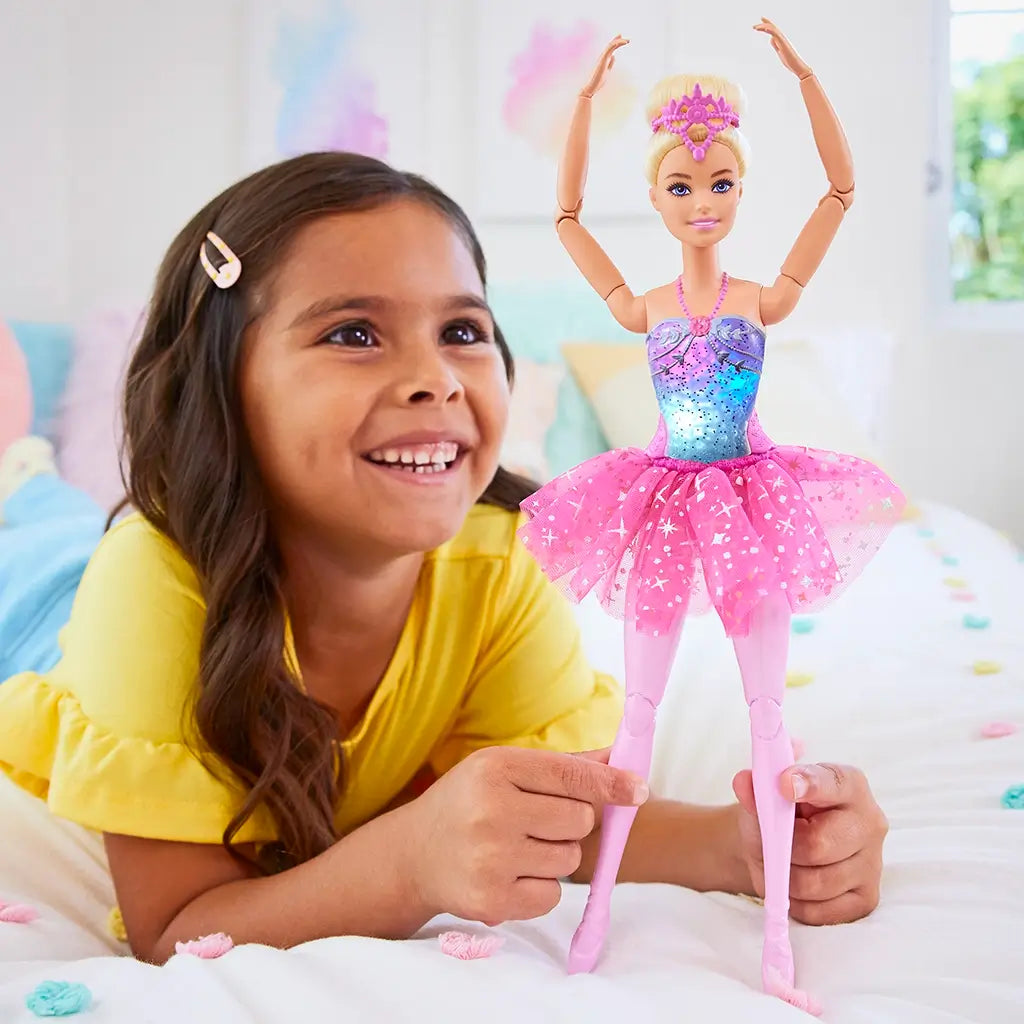 Smiling girl in yellow shirt plays with Barbie Dreamtopia Twinkle Lights Ballerina doll, dressed in a sparkling pink tutu and glowing bodice, posed in a ballet arm position.