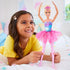 Smiling girl in yellow shirt plays with Barbie Dreamtopia Twinkle Lights Ballerina doll, dressed in a sparkling pink tutu and glowing bodice, posed in a ballet arm position.