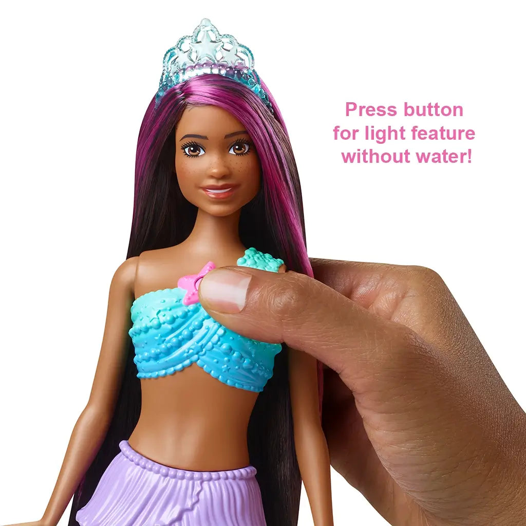 Barbie Dreamtopia Twinkle Lights Mermaid doll with brown skin, long black hair with pink streaks, wearing a blue top and tiara, shown with button feature for light-up play.