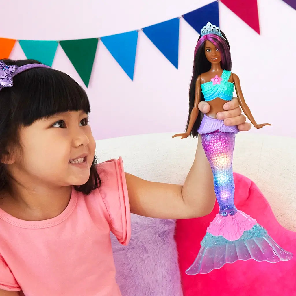 Smiling young girl in a pink shirt holds a Barbie Dreamtopia Twinkle Lights Mermaid doll with glowing rainbow tail, sitting in a bright room with colorful wall banners.