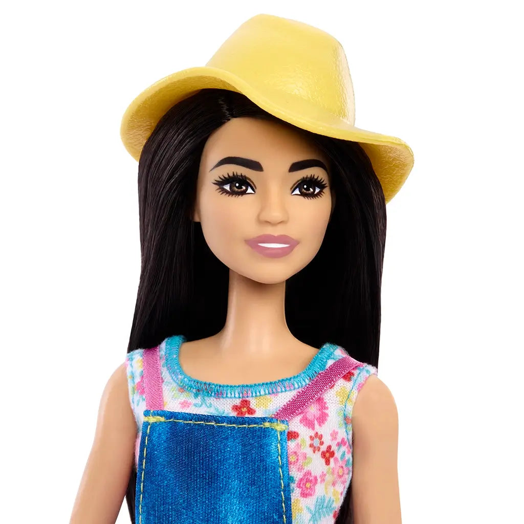 Close-up of Barbie farmer doll with long black hair, yellow hat, denim overall dress, and floral shirt, part of farm-themed playset. Detailed toy description for collectors and children ages 3 and up.
