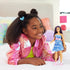 Smiling child in a shiny pink jacket plays indoors on a bed, holding Barbie Fashionistas doll dressed in a blue butterfly denim dress with pink belt and boots.