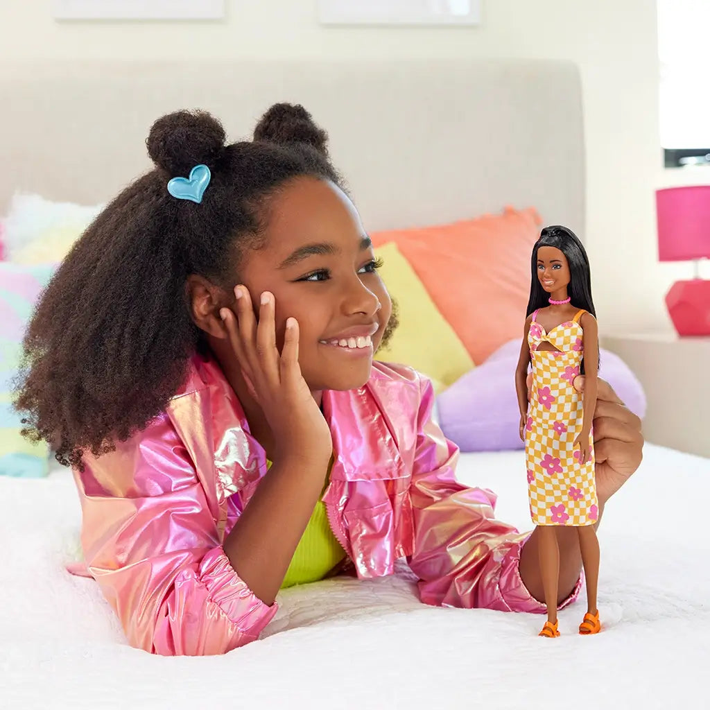 Smiling young girl plays with Barbie Fashionistas 233 doll wearing a yellow and white checkered midi dress with pink flowers and orange platform sandals.