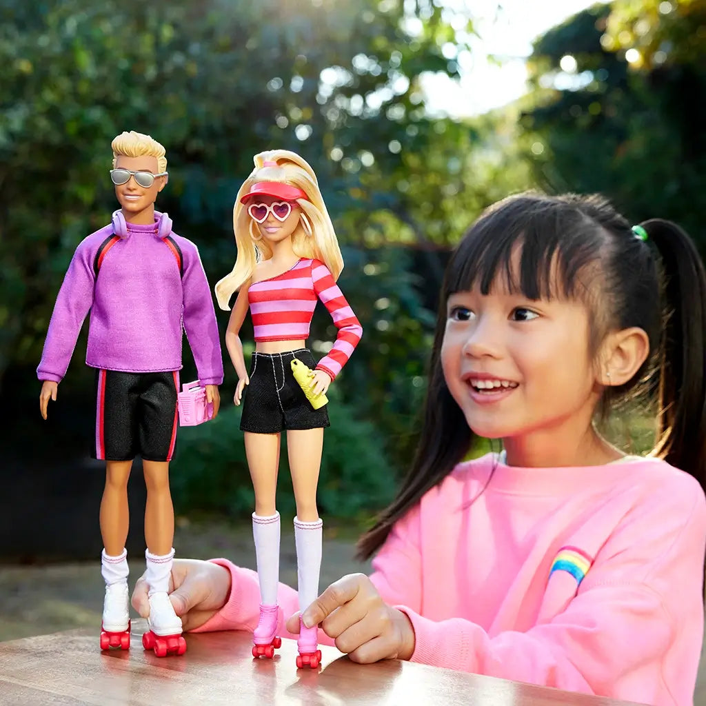 Child playing with Barbie and Ken dolls outdoors, wearing a pink sweatshirt with a rainbow design.