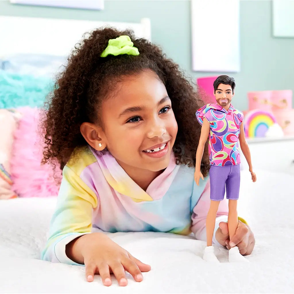 Young girl smiling while holding Barbie Fashionistas Ken doll #227 dressed in a colorful sleeveless swirl hoodie, purple shorts, and white sneakers.