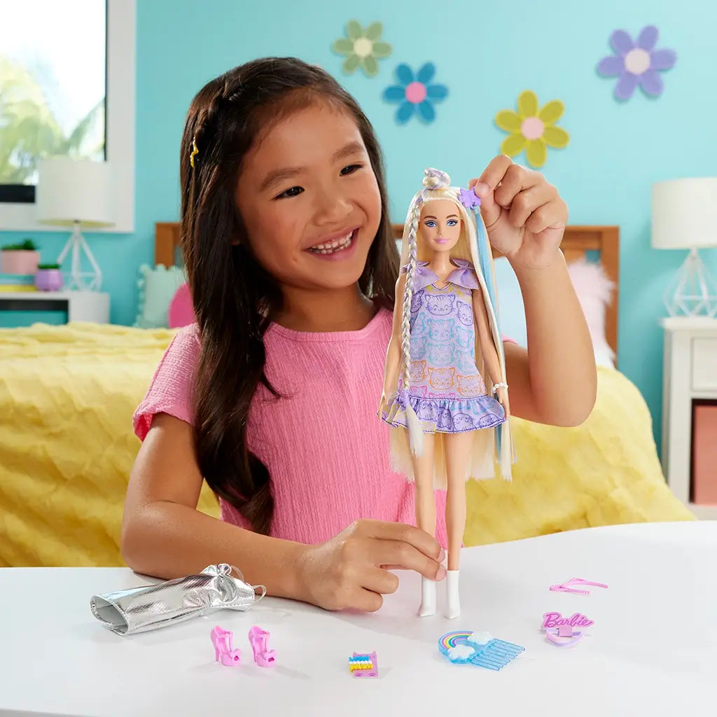 Young girl in pink dress smiling while playing with Barbie blonde fashion doll, styling its pastel kitten-print dress and arranging accessories including shoes, comb, and silver outfit.