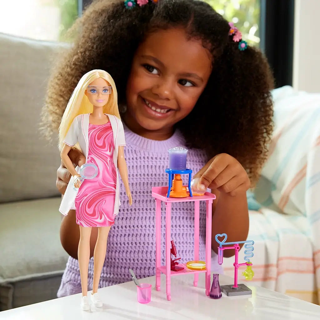 Smiling child playing with Barbie Scientist Doll dressed in pink dress and lab coat, arranging miniature lab table, microscope, and beaker accessories during fun science roleplay activity.