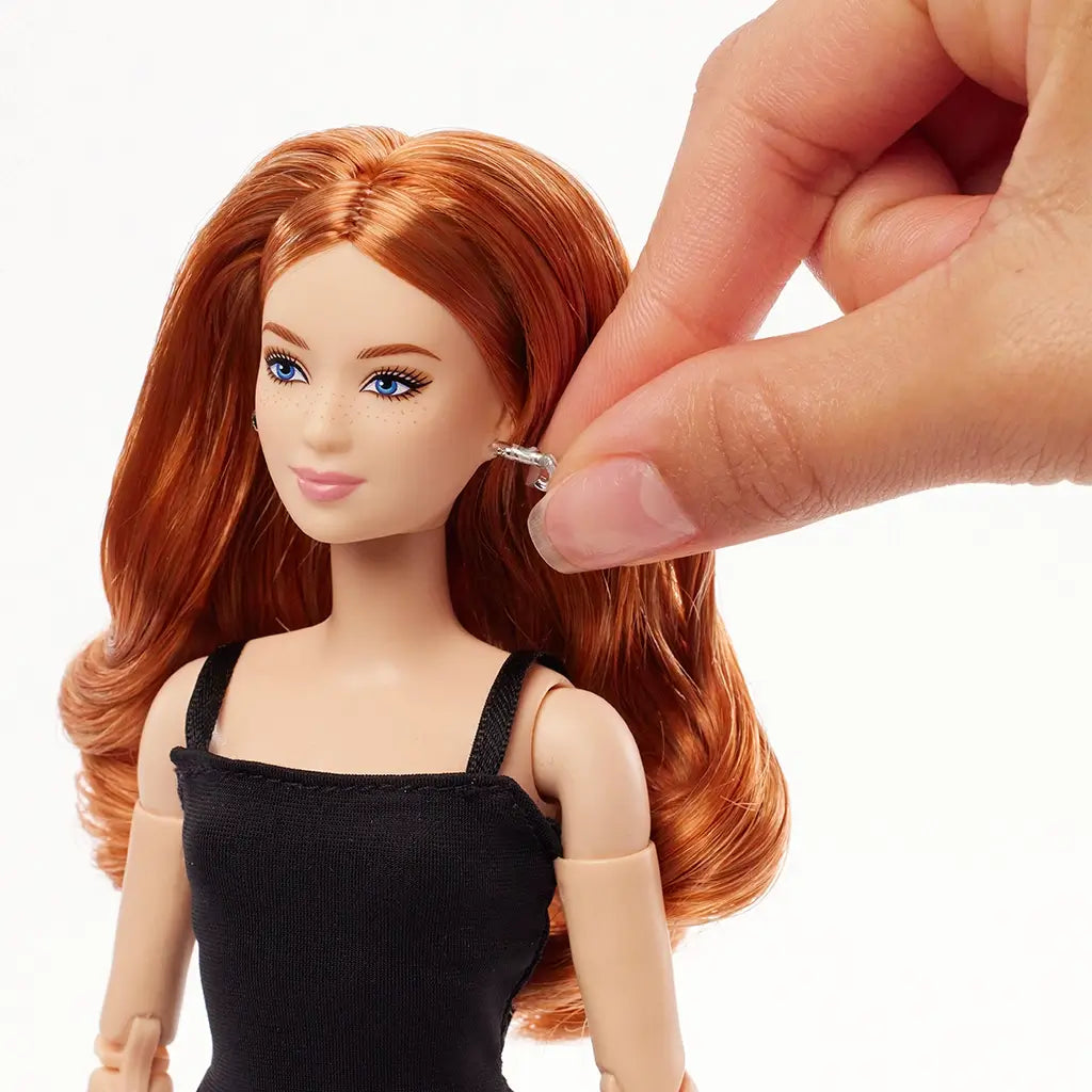 Barbie Signature Basics 2025 Model 4 fashion doll with long red hair and freckles, being fitted with removable silver hoop earring by hand while wearing black sleeveless dress.