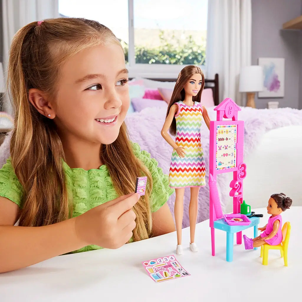 Smiling young girl plays with Barbie Teacher doll set, arranging dolls, pink chalkboard, and classroom accessories on a table in a bright, modern bedroom.