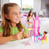 Smiling young girl plays with Barbie Teacher doll set, arranging dolls, pink chalkboard, and classroom accessories on a table in a bright, modern bedroom.