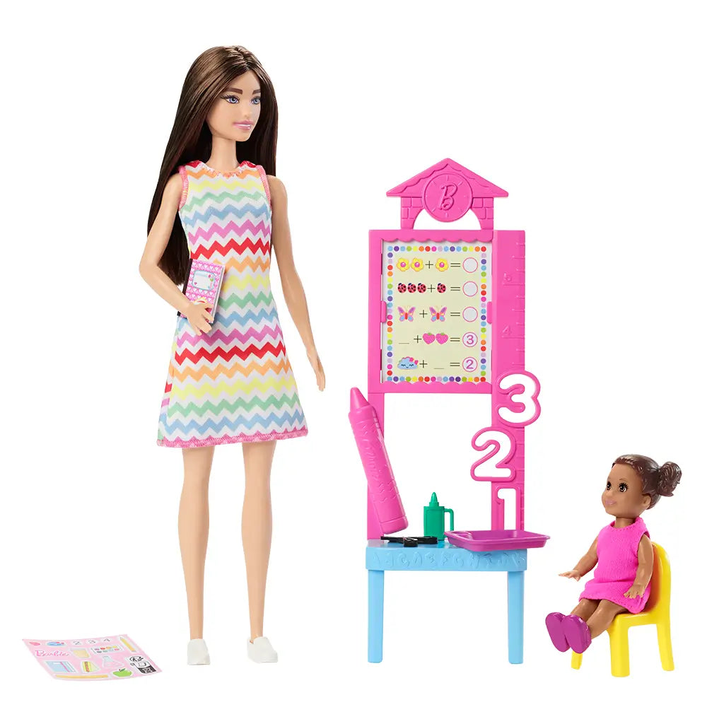 Barbie Teacher fashion doll poses beside a pink classroom board while student doll sits on a yellow chair, with miniature school supplies set on the table.