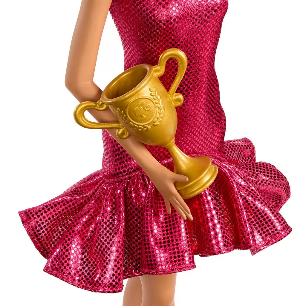 Barbie ballroom dancer doll in a glittery fuchsia one-shoulder dress holds a gold trophy with ornate handles, posed mid-dance with a flared ruffled skirt.