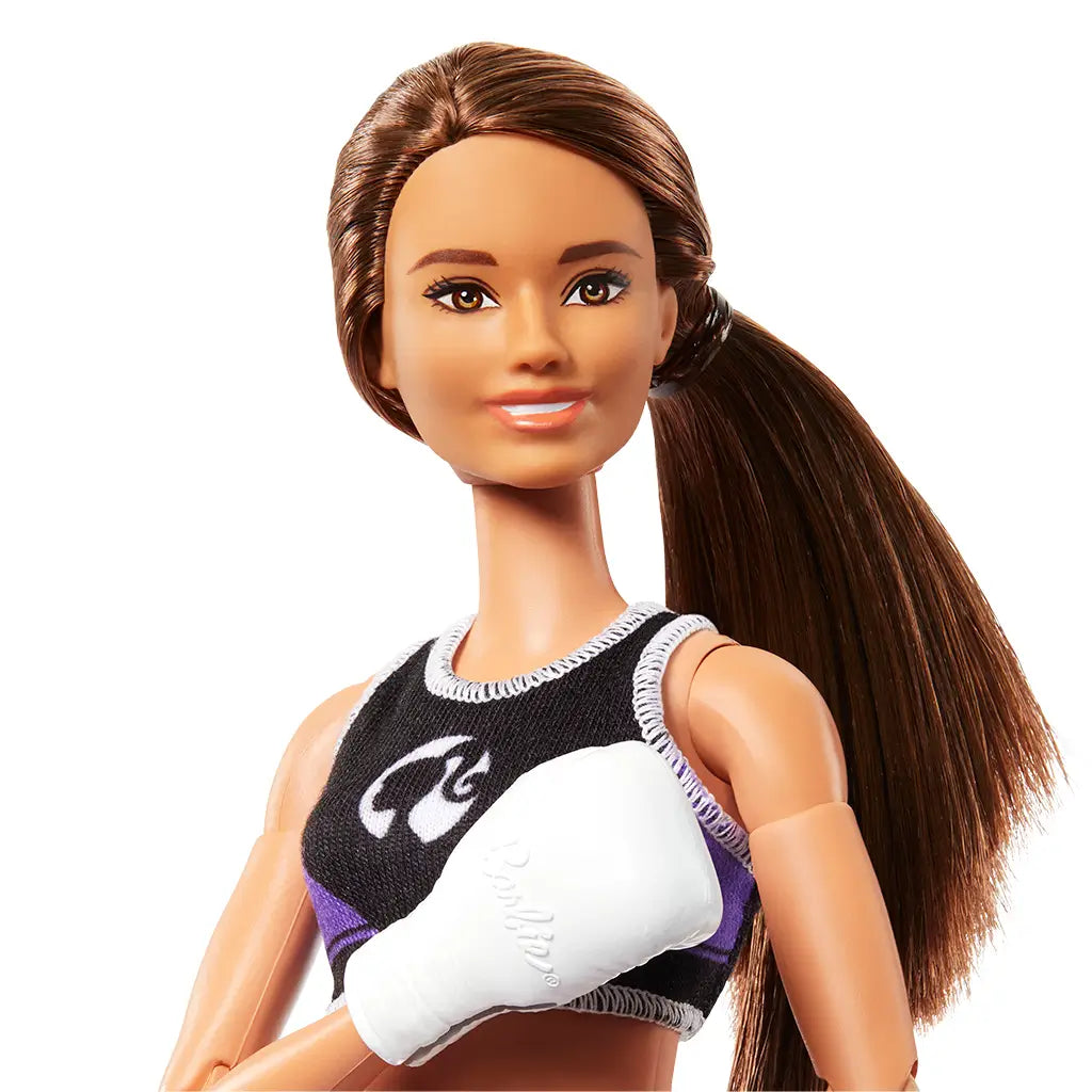 Close-up of Barbie boxer doll with long brown hair in ponytail, wearing black sports crop top with logo and white boxing gloves, smiling confidently.