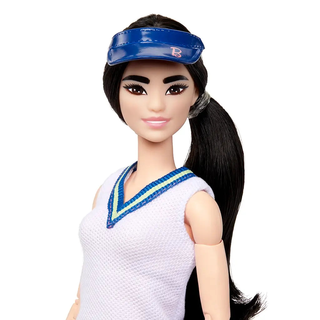Close-up of Barbie tennis player doll with long black ponytail, bright pink lips, and a shiny blue visor, wearing a white sleeveless top with blue trim neckline.